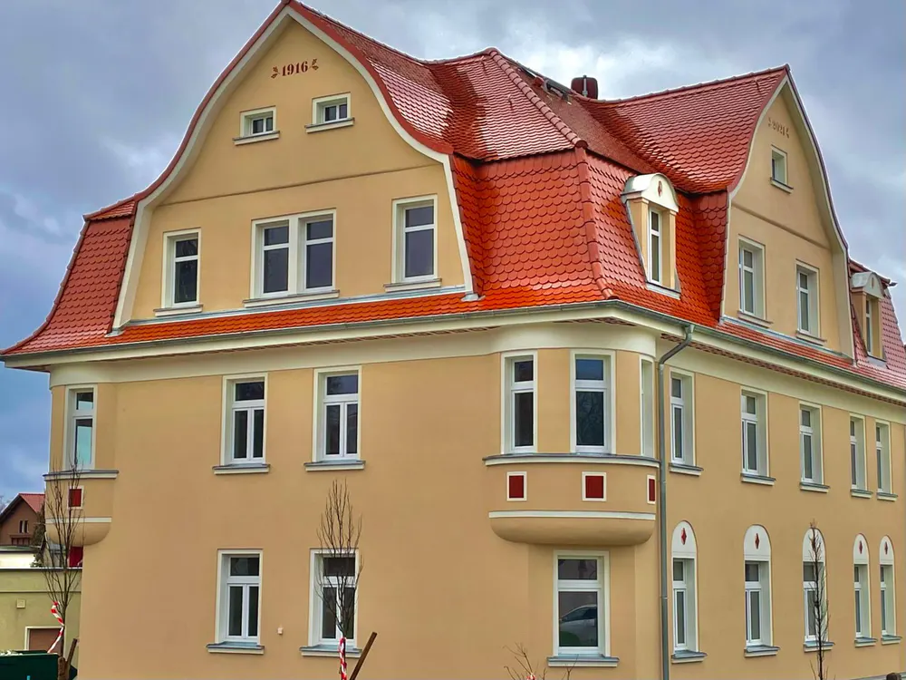 Großes, beiges Gebäude mit rotem Ziegeldach, vielen weißen Fenstern und der Aufschrift "J 1908 C" auf dem Dach.