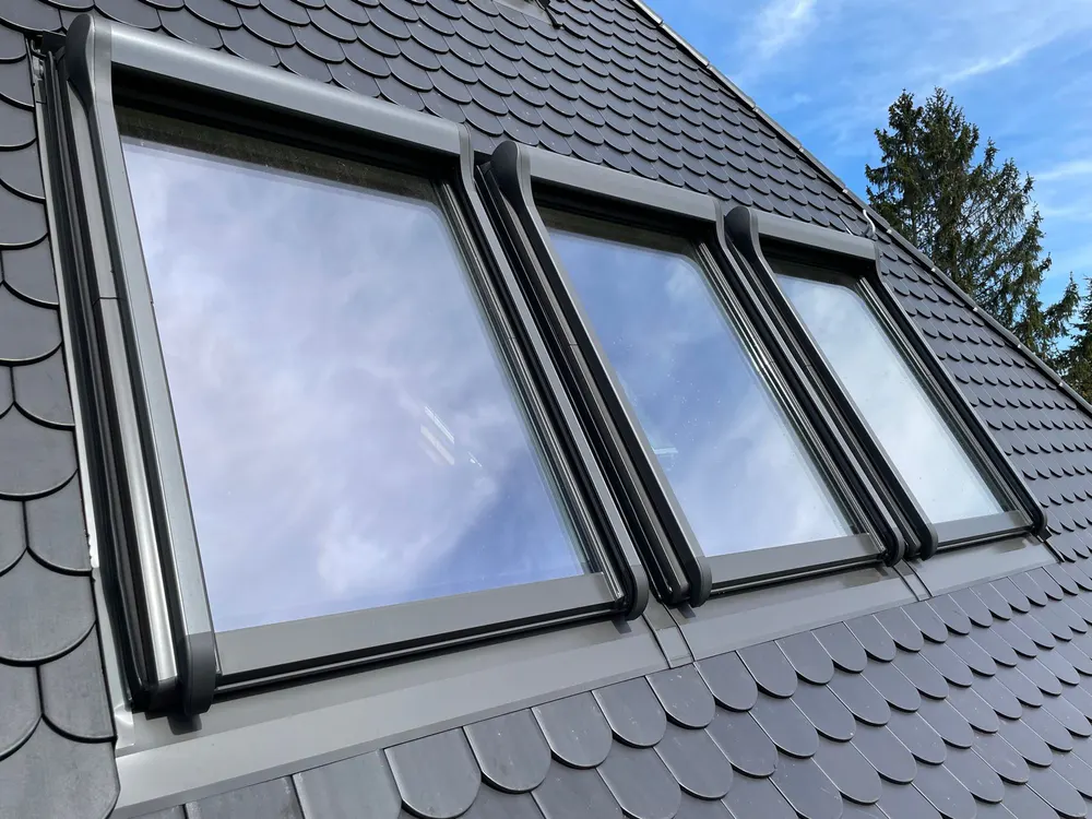 Drei Dachfenster mit grauem Rahmen in einem dunklen Schieferdach, das einen blauen Himmel widerspiegelt.