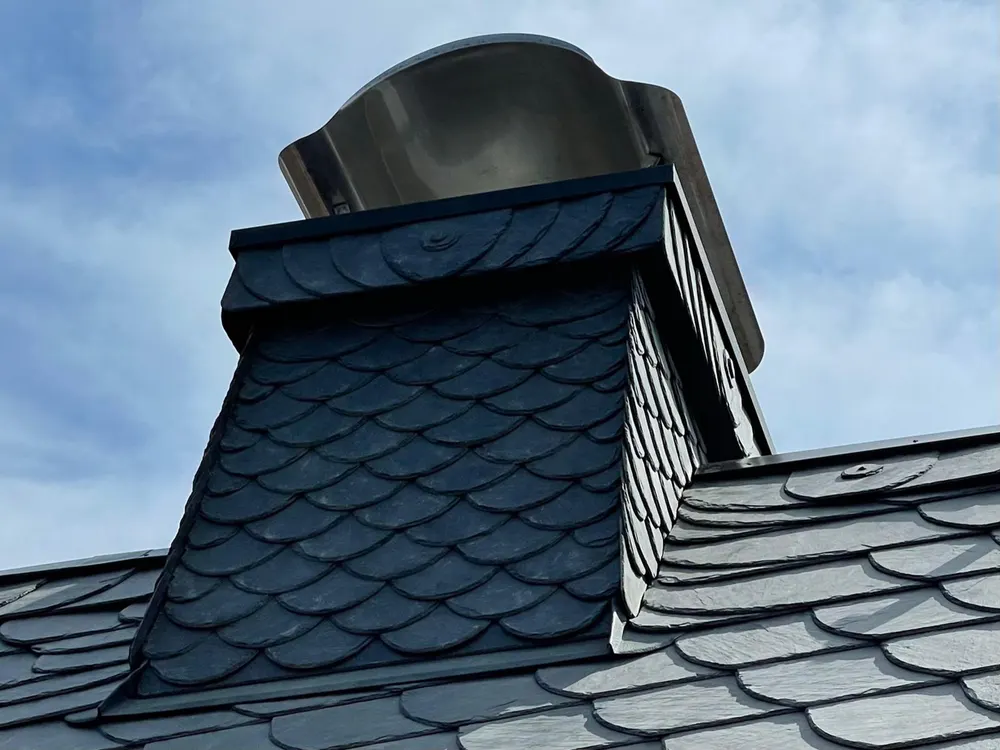 Schwarzer Schornstein mit Fischschuppen-Schieferverkleidung und glänzendem Metallaufsatz auf einem Schieferdach unter blauem Himmel.
