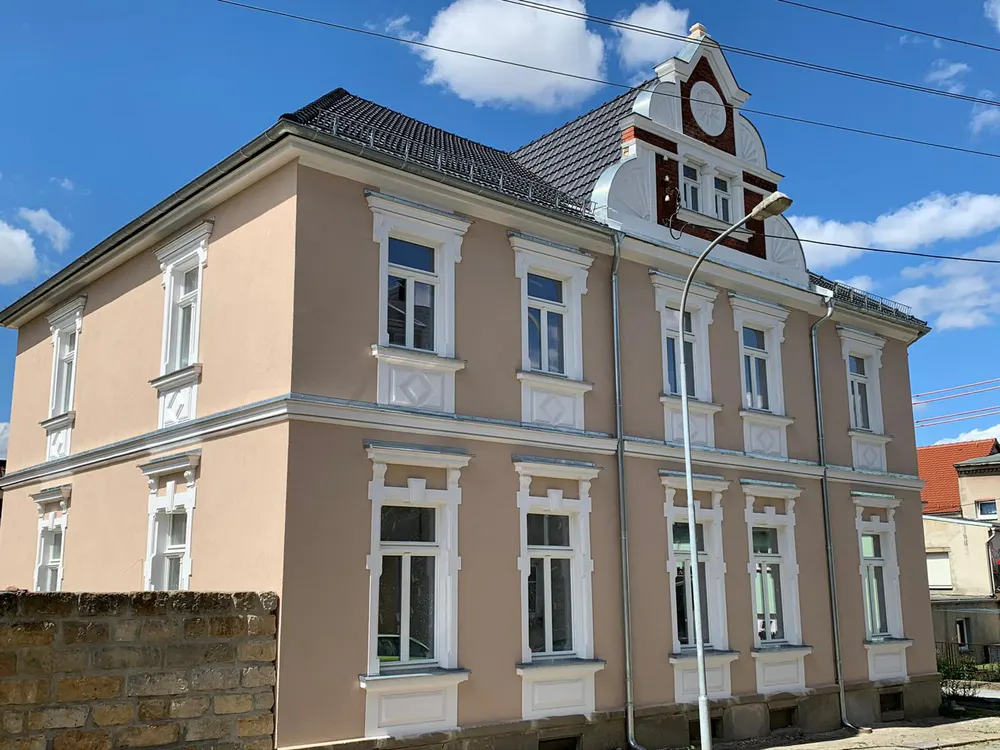 Ein großes, hellrosa Mehrfamilienhaus mit weißen Fensterrahmen und Dachgiebeln unter blauem Himmel.