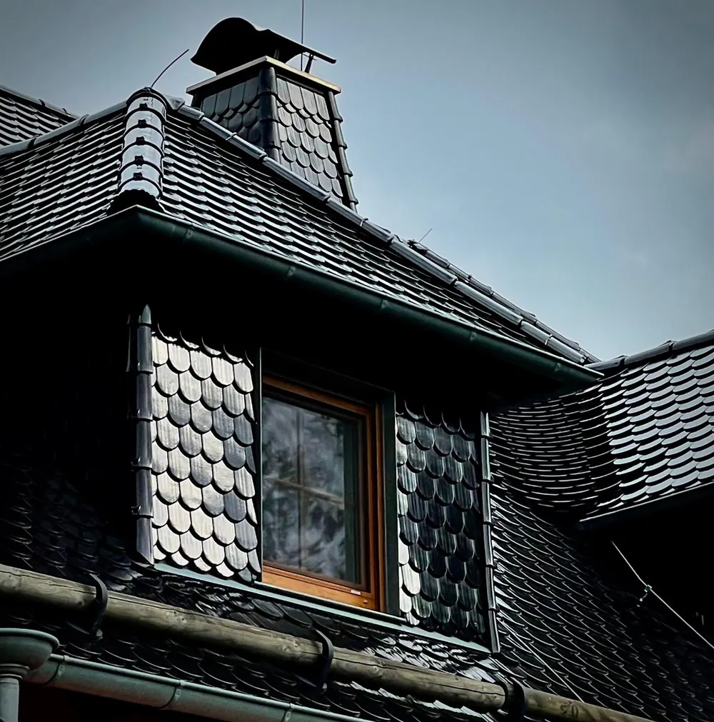 Dach mit Schieferziegeln, Gaube, Holzfenster, Dachrinne und Schornstein unter bewölktem Himmel.