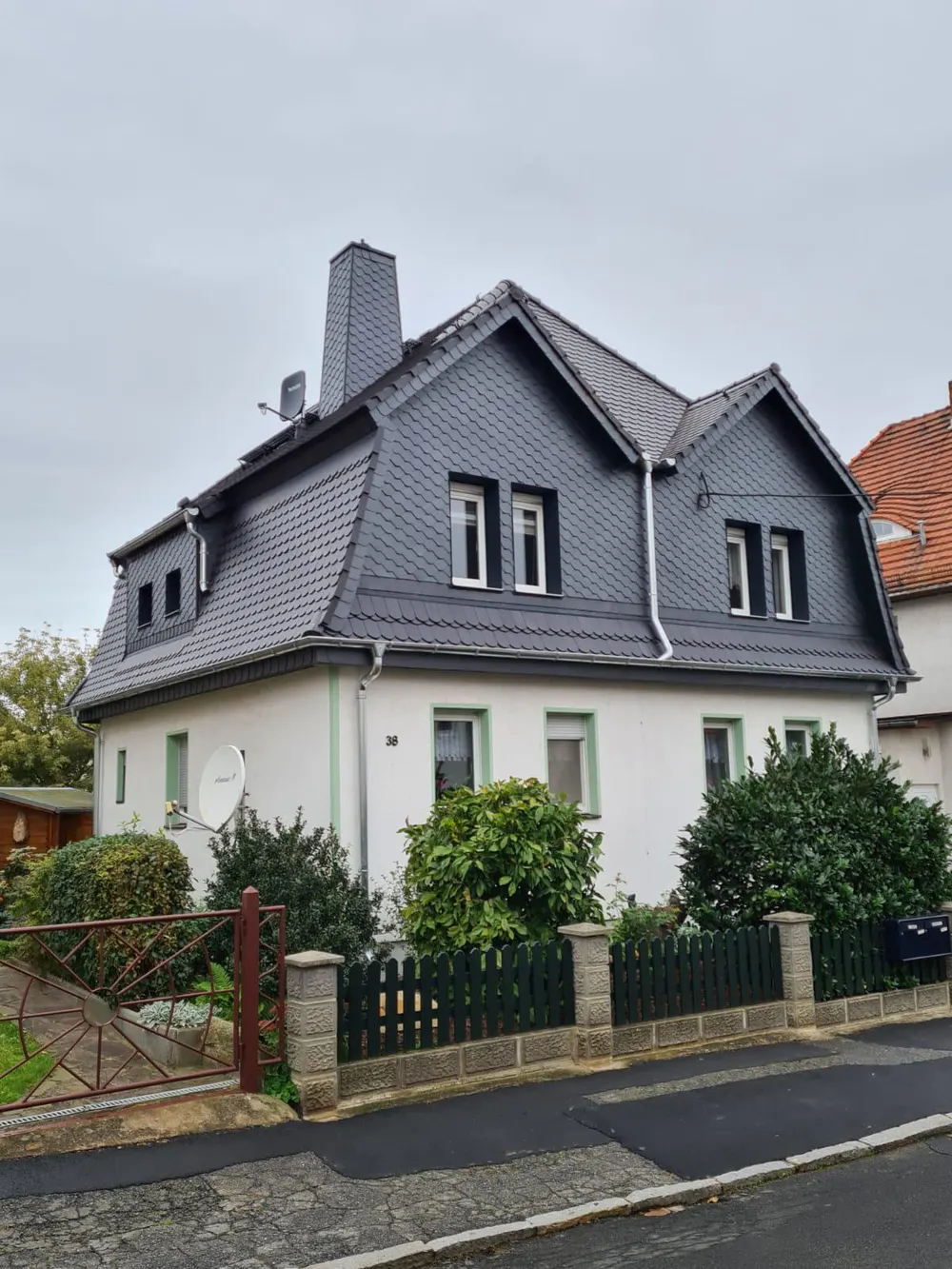 Weißes Haus mit dunkelgrauem Schieferdach, Schornstein, Dachgauben und grünem Holzzaun.