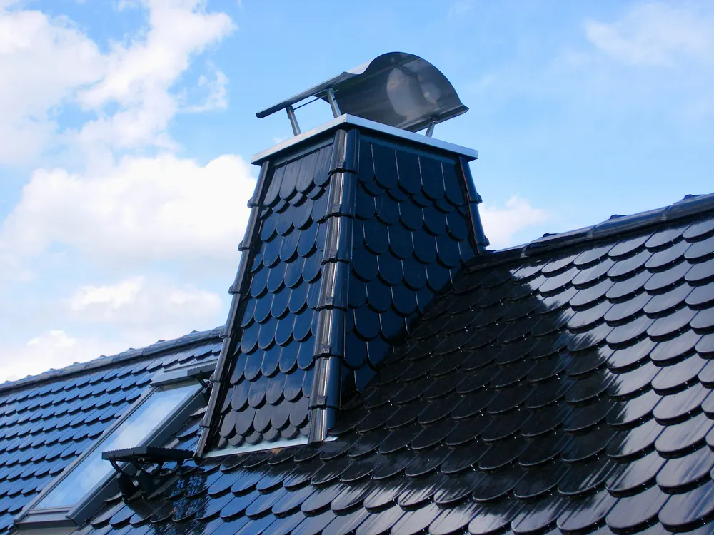 Schwarzer Schornstein mit glänzenden Schindeln auf einem Dach mit Dachfenstern unter blauem Himmel.