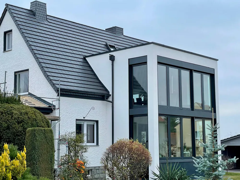 Modernes weißes Haus mit dunklem Dach und Glasanbau, umgeben von grünen Büschen.